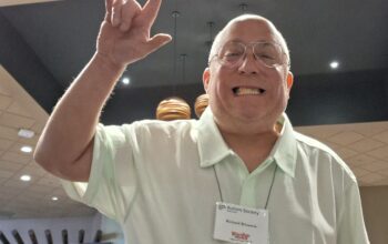 A white guy with a white short-sleeved short waves a hand.