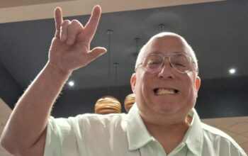 A white man with glasses holds his hand up.