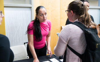 A BIPOC woman with long black hair wearing a pink T-shirt stands at a table to talk to a young white woman with a backpack.