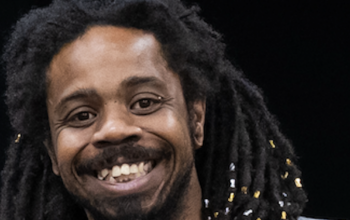 A Black man with a mustache and beard and long black hair smiles in a headshot.