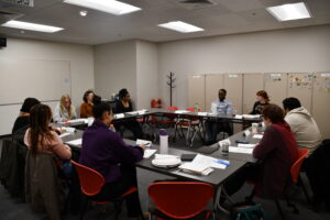 A group of people of diverse identities sit around a table at a listening session.