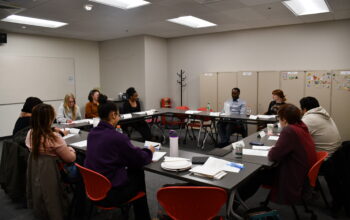 A group of people of diverse identities sit around a table at a listening session.