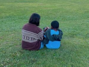 The backs of two people -- one a young woman in a purple shirt and the other a small child in a blue shirt -- sitting on a green lawn.