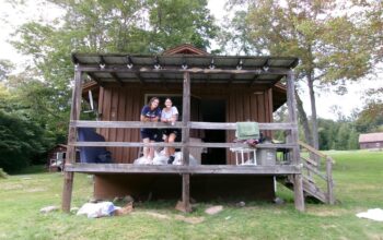 A picture of the outdoors with green trees and a cabin at a summer camp with two people at the railing looking down.