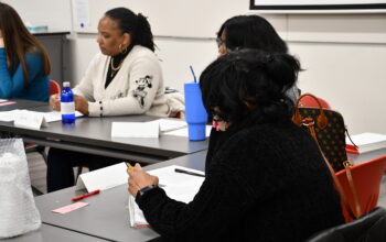 A Black woman with a ponytail talks to a group at a table.