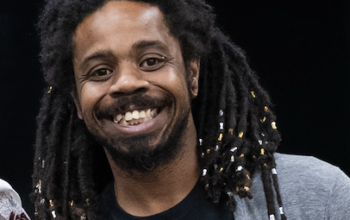 A Black man with a mustache and beard and long black hair smiles in a headshot.