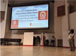 A Black woman stands on a stage holding a mic with a large screen behind her that says collaborative storytelling class.