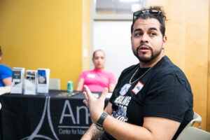 A BIPOC man with black hair is gesturing in conversation in a room with yellow walls.